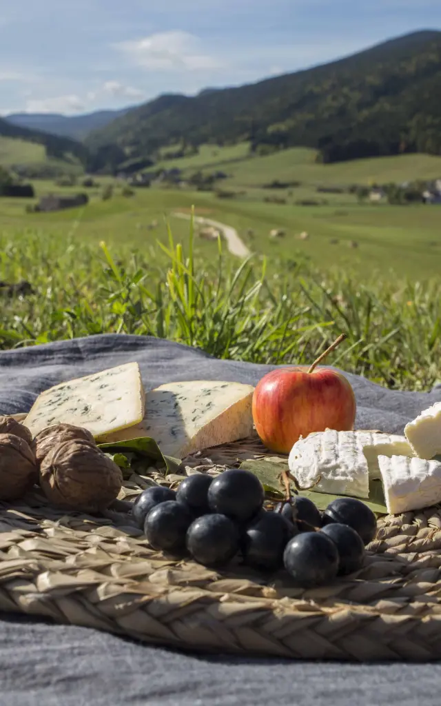 Panier en osier rempli de produits gourmands : fromages, charcuterie, fruits frais et pain, posé sur une couverture en tissu à motifs