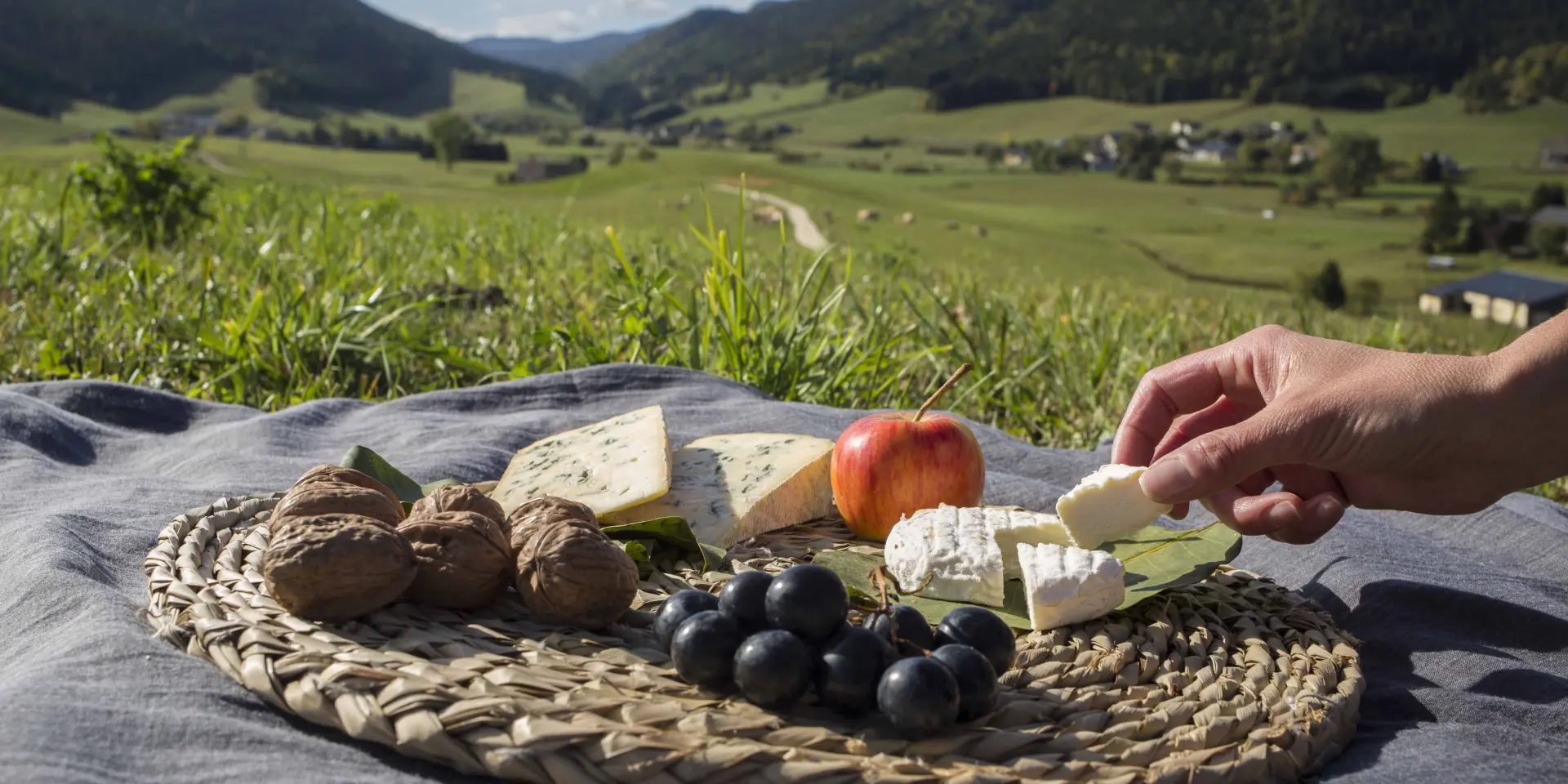Panier en osier rempli de produits gourmands : fromages, charcuterie, fruits frais et pain, posé sur une couverture en tissu à motifs