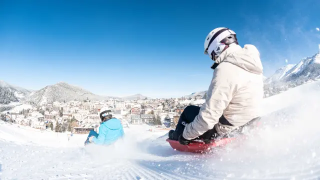 Personne en combinaison de surf debout sur une planche rouge, sur une vague artificielle enneigée avec des montagnes en arrière-plan