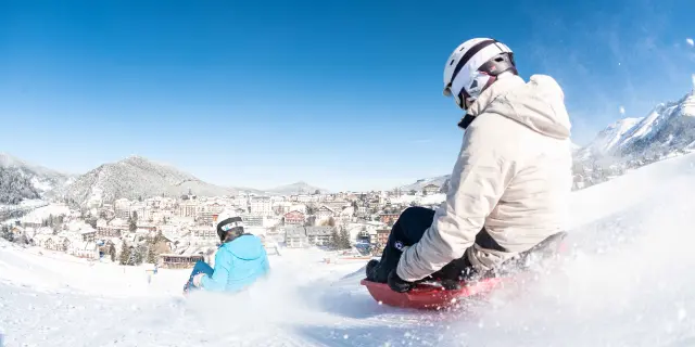 Personne en combinaison de surf debout sur une planche rouge, sur une vague artificielle enneigée avec des montagnes en arrière-plan