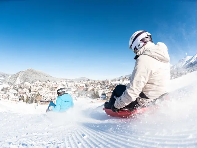 Persona in muta da surf in piedi su una tavola rossa, su un'onda artificiale innevata con montagne sullo sfondo