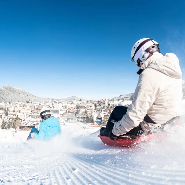 Personne en combinaison de surf debout sur une planche rouge, sur une vague artificielle enneigée avec des montagnes en arrière-plan