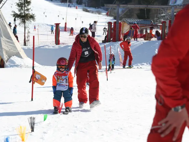 Due bambini in tuta da sci rossa e blu, con racchette e sci, su una pista da sci soleggiata