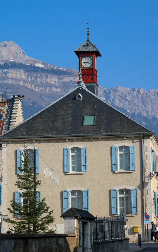 Torre dell'orologio in legno con un orologio rosso e un tetto in tegole, circondata da montagne innevate