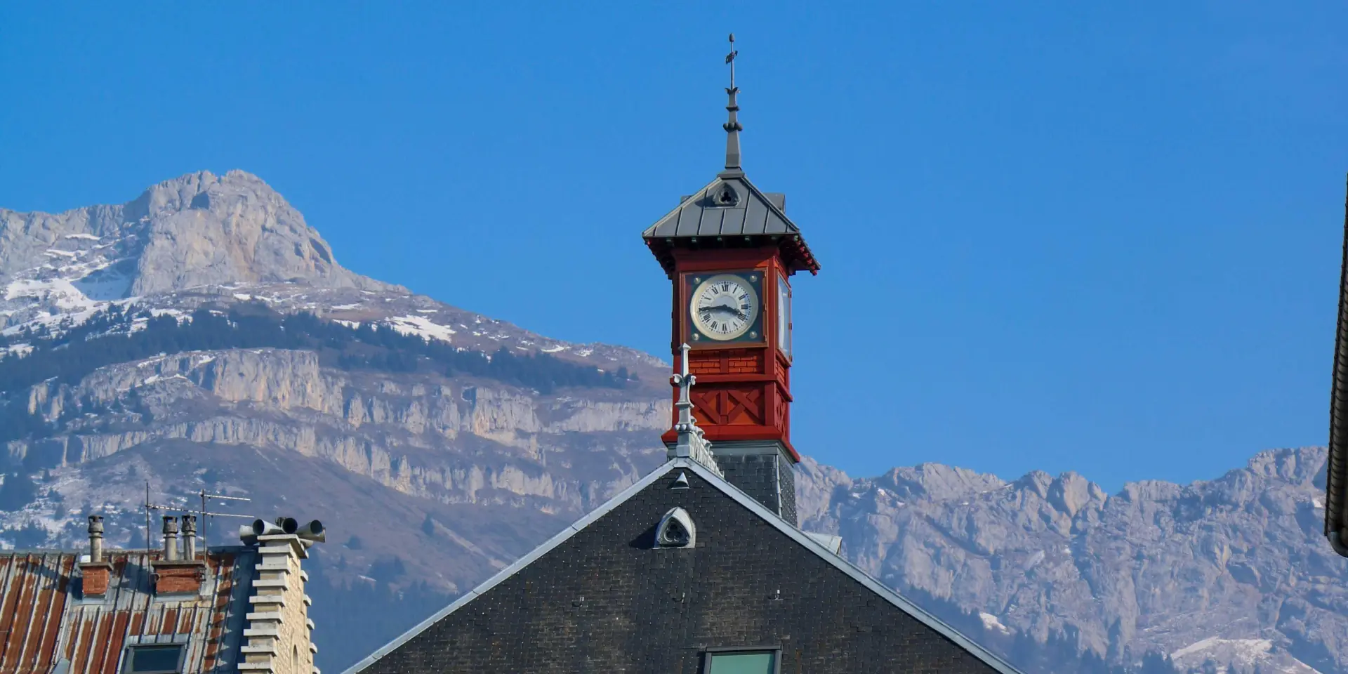 Torre dell'orologio in legno con un orologio rosso e un tetto in tegole, circondata da montagne innevate