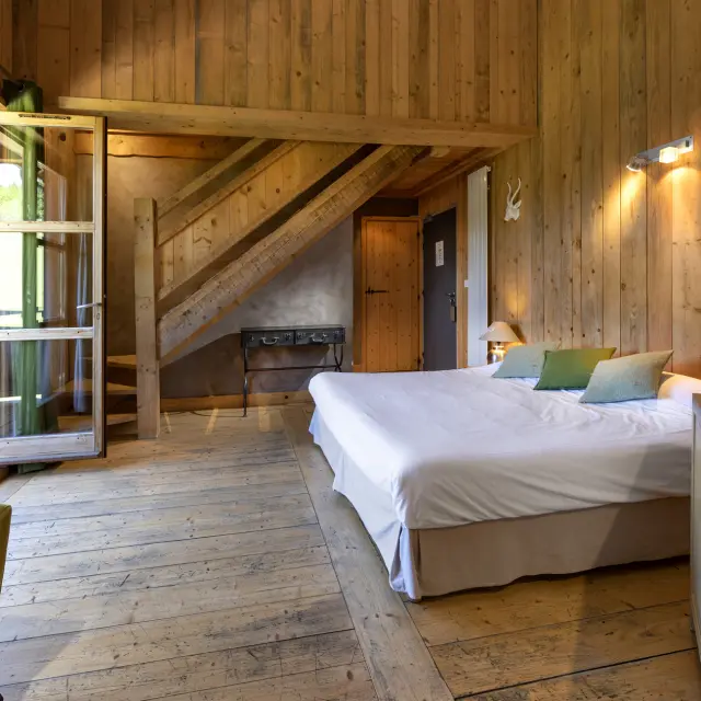 Chambre d’hôtel avec lit double, tête de lit en bois, table de chevet et fenêtre donnant sur un balcon