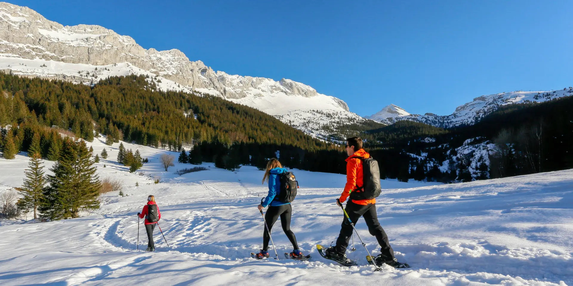 Tre persone in abbigliamento da sci di fondo che attraversano un lago ghiacciato innevato, con montagne sullo sfondo