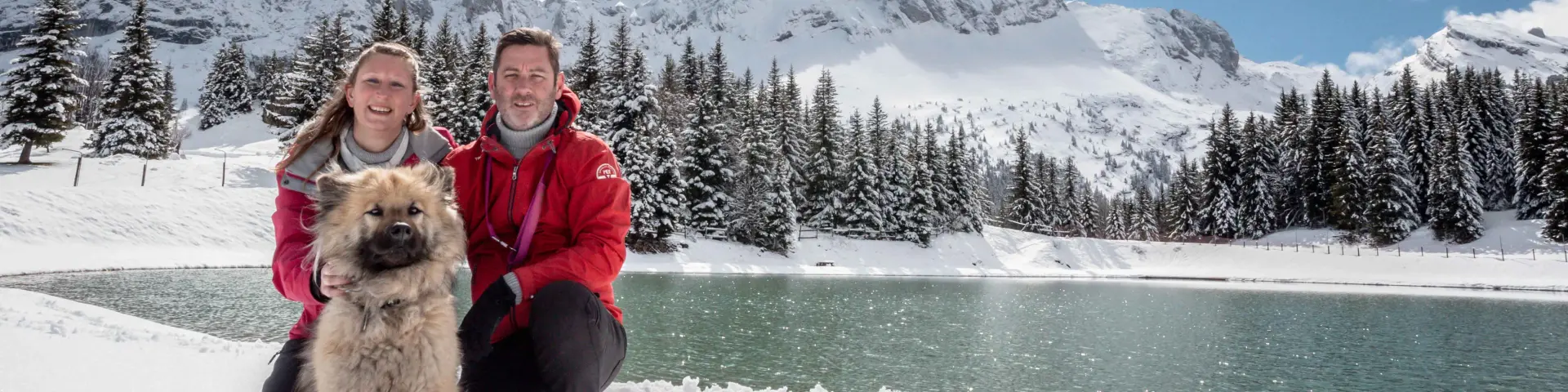 Deux personnes en tenue de ski tenant un husky en laisse, sur un sentier enneigé avec des montagnes en arrière-plan