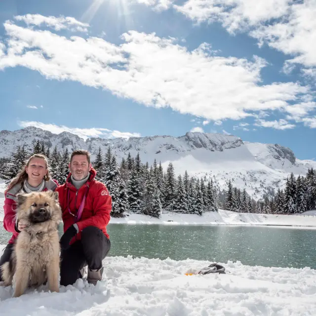 Deux personnes en tenue de ski tenant un husky en laisse, sur un sentier enneigé avec des montagnes en arrière-plan