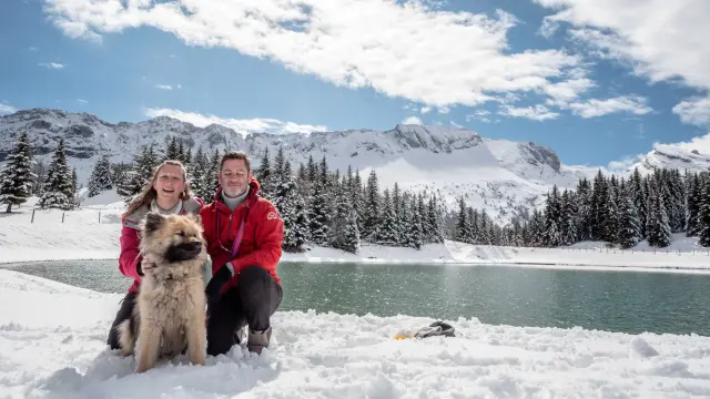 Due adulti e un bambino in abbigliamento invernale con un cane husky di fronte a un lago ghiacciato e montagne innevate