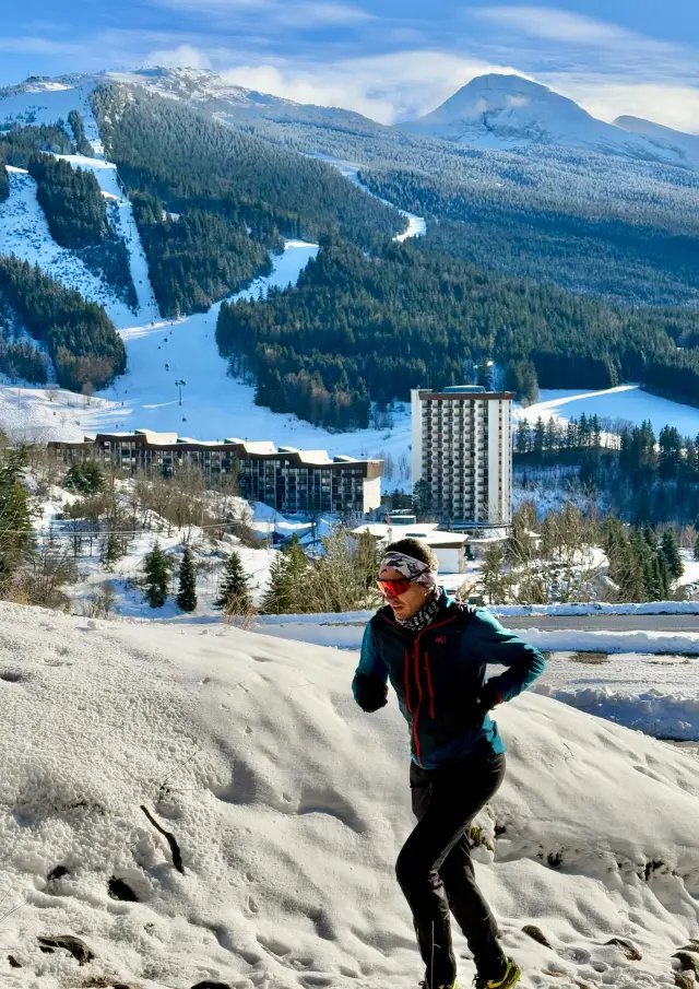 Person in hiking gear walking on a snowy trail with a lake and mountains in the background