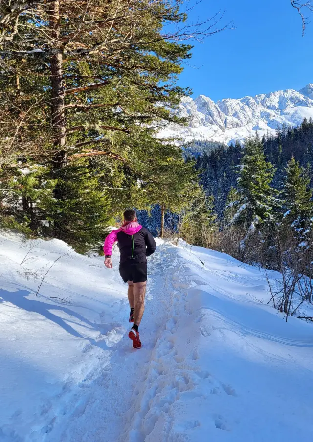 Two people walking on a snowy trail in the mountains, with trees and snow-capped peaks in the background