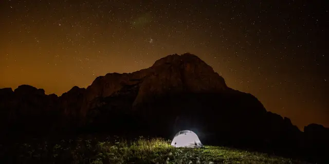 Paysage nocturne montrant une montagne sombre et étoilée avec une tente éclairée en contrebas