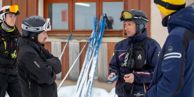 Deux secouristes en combinaison de protection bleue et jaune, dont un avec un casque jaune, sur un bateau. Un troisième secouriste en tenue noire et casque bleu les observe.