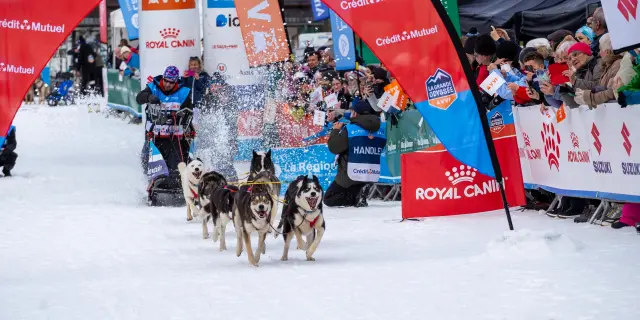 Deux skieurs de fond en compétition sur une piste éclairée, avec public et sponsors en arrière-plan