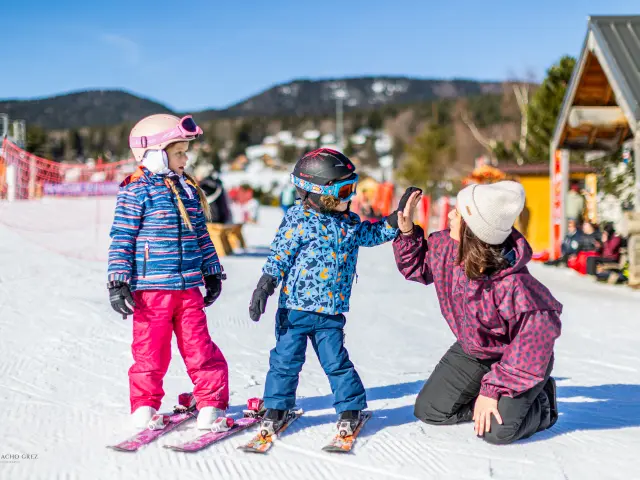 Trois personnes en tenue de ski, dont deux enfants, sur une piste enneigée avec des bâtiments et des montagnes en arrière-plan